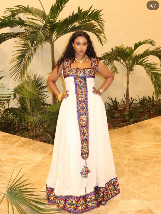 Woman wearing a white sleeveless Habesha kemis featuring colorful geometric Tilf embroidery along the square neckline, vertical front panel, and hem.