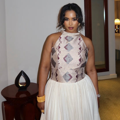 A woman wearing a sleeveless halter-neck Ethiopia clothes with a textured beige and burgundy geometric pattern on the bodice and a flowing white skirt, standing indoors.