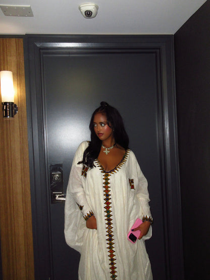 A woman wearing a white long-sleeve traditional Ethiopian Dress  with brown and gold geometric embroidery, standing in front of a dark gray door.