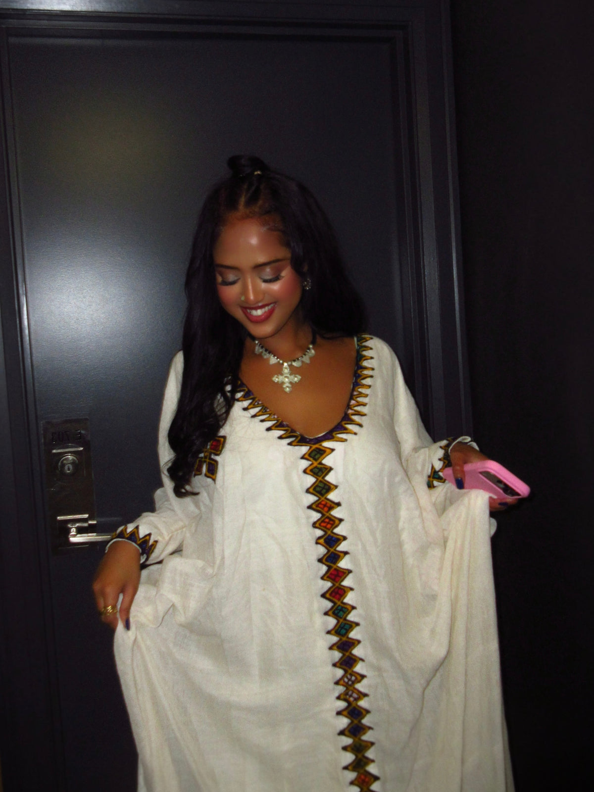 A woman wearing a white long-sleeve Ethiopian Dress  with colorful geometric embroidery down the front and neckline, standing in front of a dark door.