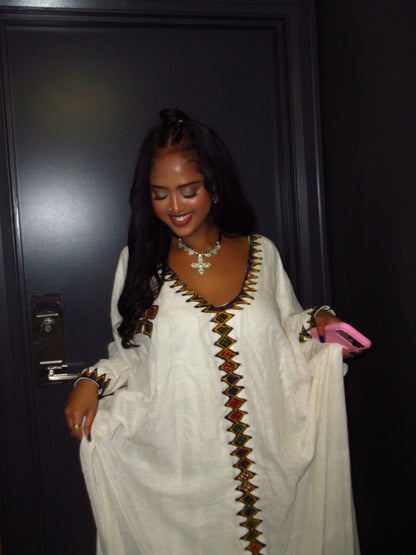 A woman wearing a white long-sleeve Ethiopian Dress  with colorful geometric embroidery down the front and neckline, standing in front of a dark door.