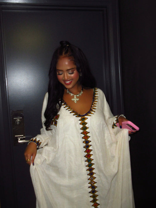 A woman wearing a white long-sleeve Ethiopian Dress  with colorful geometric embroidery down the front and neckline, standing in front of a dark door.