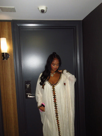 A woman wearing a white Ethiopian Dress  with brown and gold geometric embroidery, standing in front of a dark gray door in a hotel hallway.