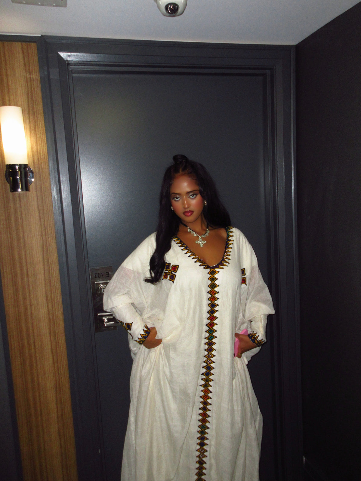 A woman wearing a white long-sleeve Ethiopian Dress  with brown geometric embroidery stands in front of a dark gray door.