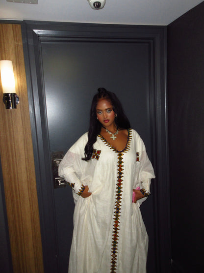 A woman wearing a white long-sleeve Ethiopian Dress  with brown geometric embroidery stands in front of a dark gray door.