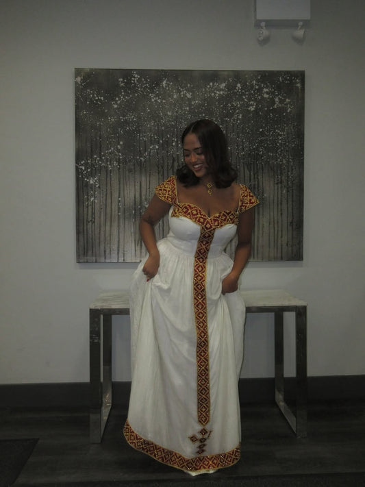 Woman in a white and patterned Habesha kemis standing in a room with a painting on the wall.