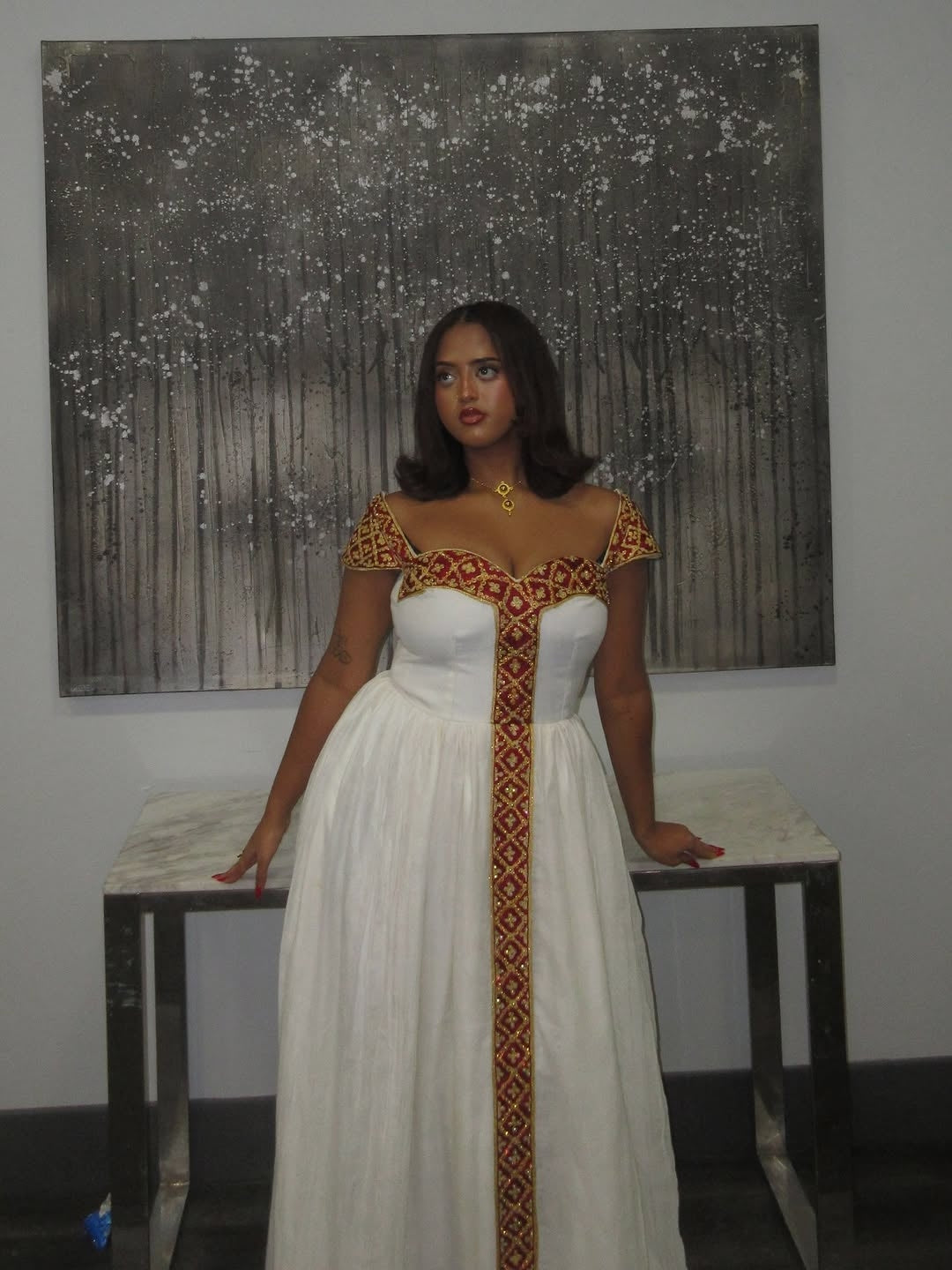 Woman in a white habesha kemis with decorative patterns standing in front of a painting.
