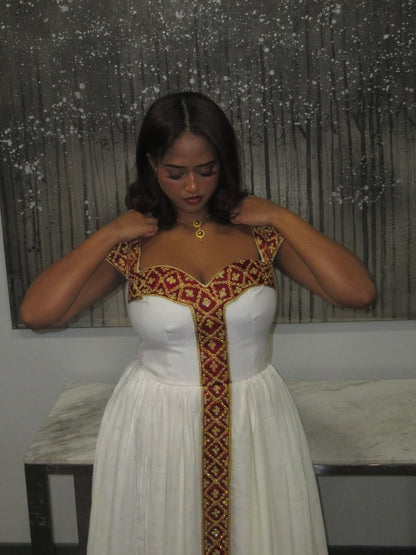 Woman wearing a white habesha kemis with red and gold embroidery in a room.