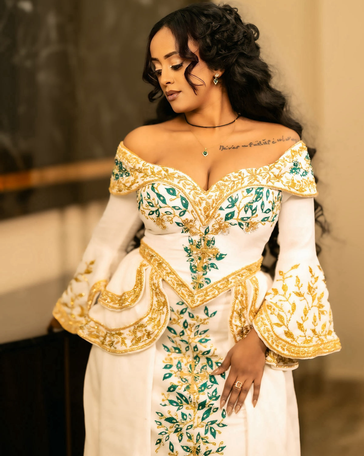 A woman wearing an off-the-shoulder white habesha Wedding dress with ornate gold and green embroidered detailing, posed indoors against a softly blurred background.