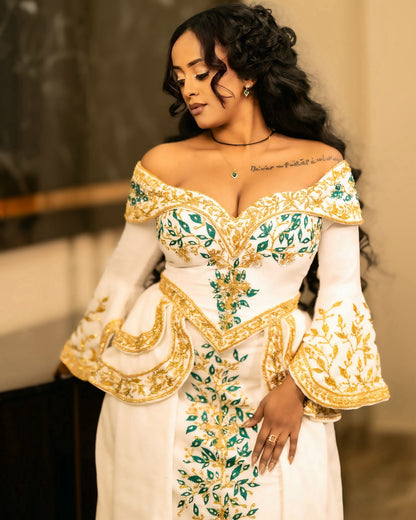 A woman wearing an off-the-shoulder white habesha Wedding dress with ornate gold and green embroidered detailing, posed indoors against a softly blurred background.