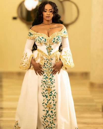 A woman modeling an off-the-shoulder white habesha Wedding dress with ornate gold embroidery and green floral detailing, standing in a warmly lit indoor setting.