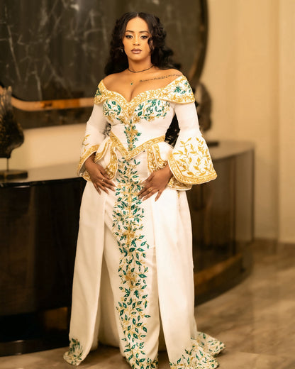 A woman models a luxurious off-the-shoulder white habesha Wedding dress with elaborate gold embroidery and green floral patterns, set against an elegant indoor background.