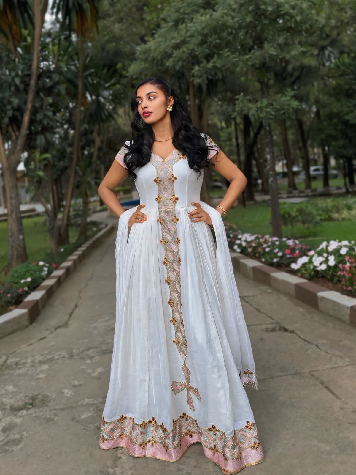 A woman stands outdoors on a paved path in a park, wearing a long white traditional Habesha Zuria with gold embroidery and a pink border at the hem.