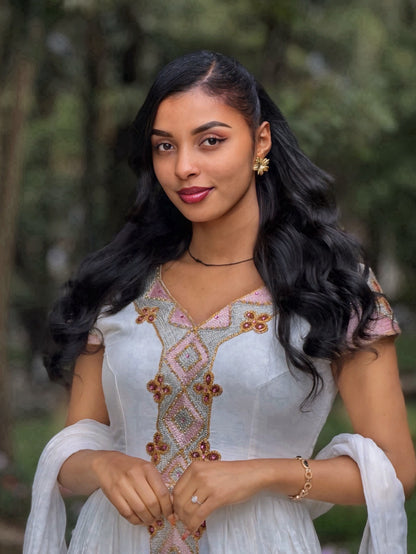A woman with long, wavy black hair wearing a white traditional Habesha Zuria with pink and gold embroidery, standing outdoors against a blurred green background.