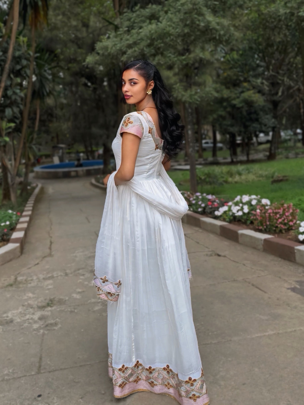 A woman wearing a white traditional Habesha Zuria with floral embroidery stands on a paved path in a park, holding the side of the dress to show its drape.