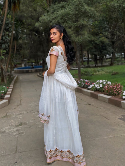 A woman wearing a white traditional Habesha Zuria with floral embroidery stands on a paved path in a park, holding the side of the dress to show its drape.