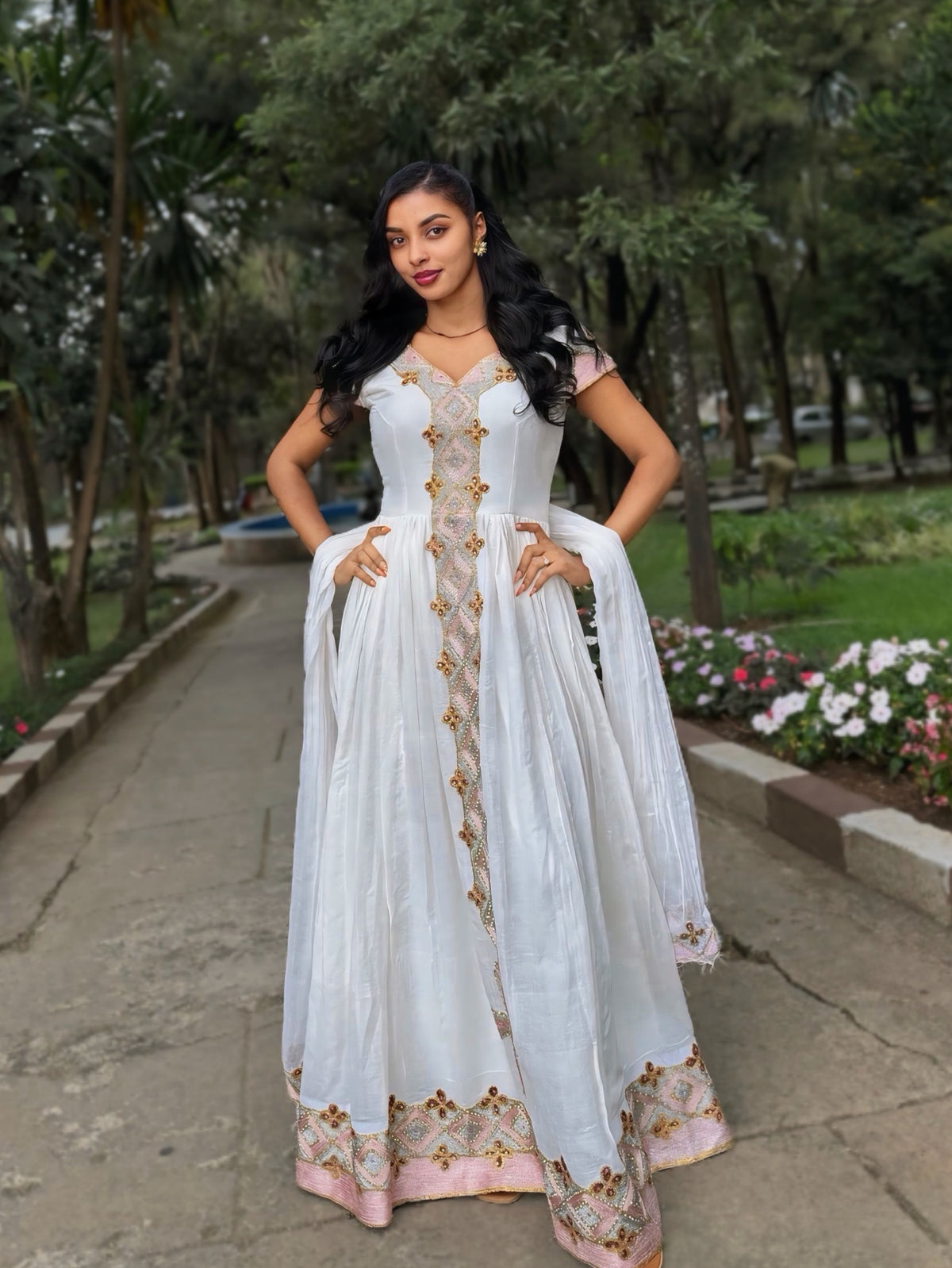 A woman stands outdoors on a paved path in a park, wearing a white traditional Habesha Zuria with gold and pink floral embroidery and a matching sheer dupatta.