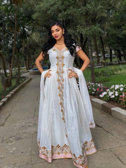 A woman stands outdoors on a paved path in a park, wearing a white traditional Habesha Zuria with gold and pink floral embroidery and a matching sheer dupatta.