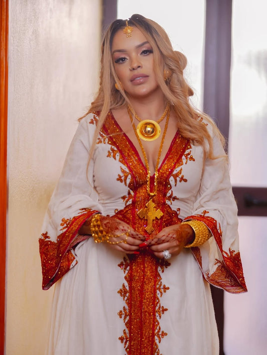 Woman wearing a white and orange embroidered Habesha Zuria with gold jewelry.