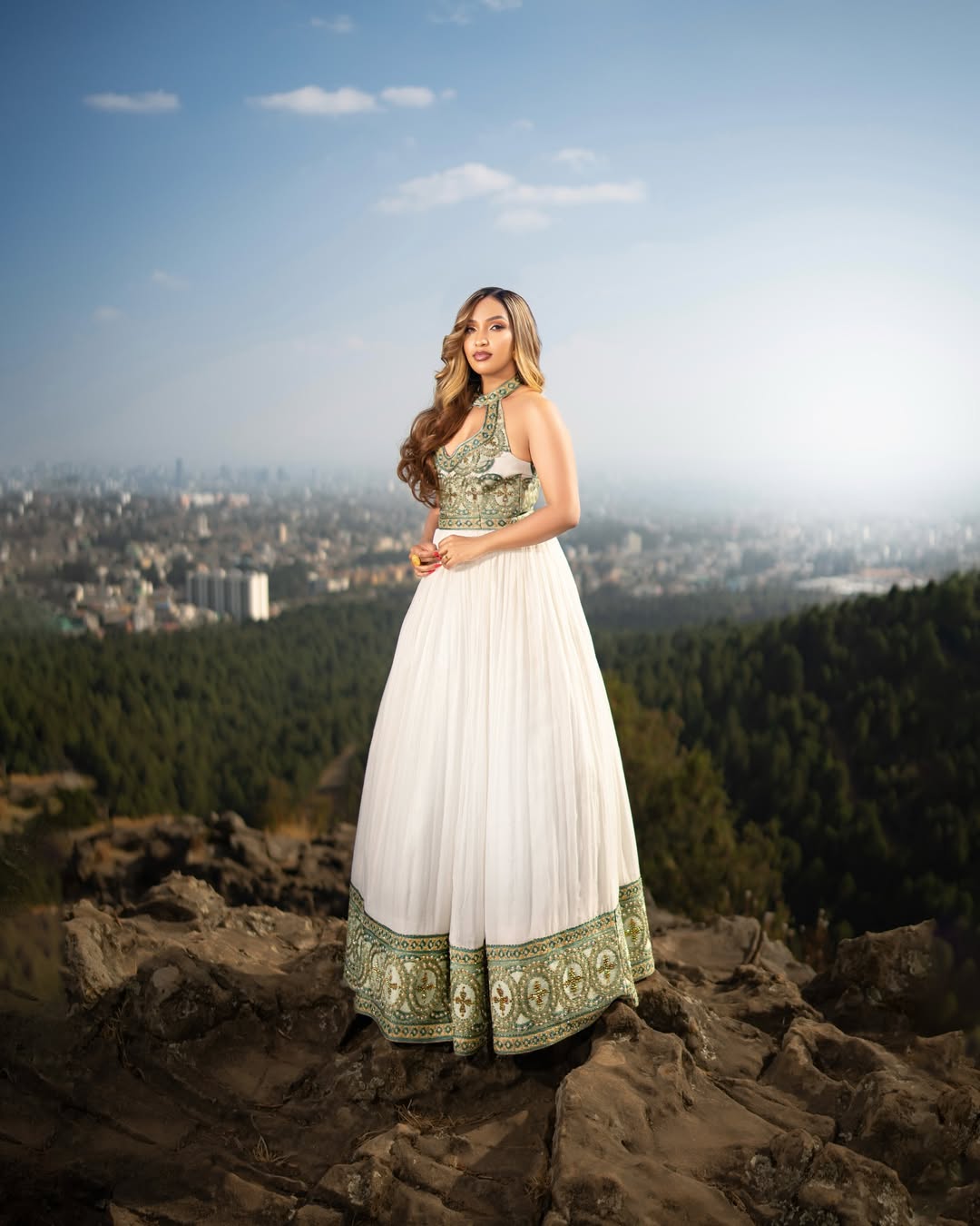 Woman in a white and green Ethiopian dress standing on a rocky outcrop with a cityscape and trees in the background