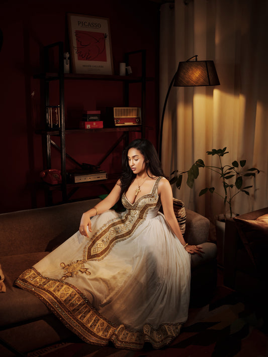 A woman sits on a sofa in a dimly lit room wearing a cream and gold embellished traditional  Habesha Kemis. A floor lamp casts warm light on the curtains, and shelves with books and framed art are visible in the background.