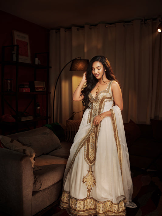 A woman wearing a white traditional  Habesha Kemis with intricate gold embroidery and a matching dupatta, standing in a warmly lit living room.