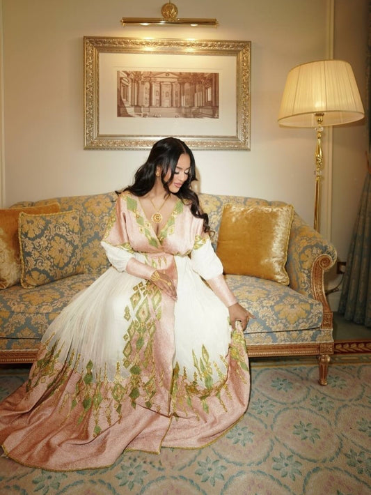 A woman wearing a traditional embroidered Ethiopian Traditional Clothing sits on an ornate sofa in a decorated room. The gown is cream and blush pink with green and gold embroidery, and the background features a patterned sofa, framed wall art, and a floor lamp.
