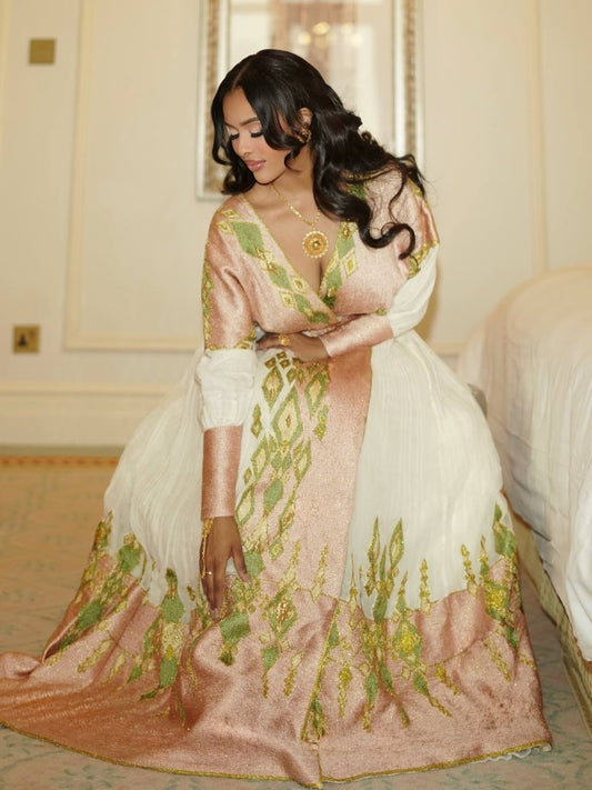 A woman wearing a traditional South Asian Ethiopian Traditional Clothing in a warm peach-pink and white fabric with green and gold embroidery, posed in an elegant indoor setting.