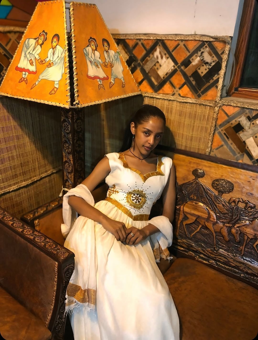 Woman in a white Ethiopian dress sitting on an ornate wooden bench with decorative lampshades.