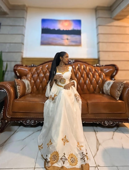 Woman in a white and gold dress sitting on a brown leather couch in a room with a sunset painting on the wall.
