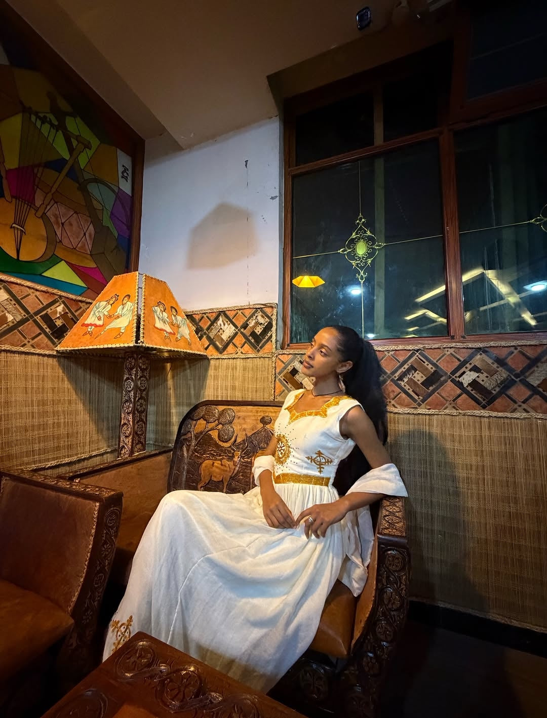 Woman in a white Ethiopian dress sitting in a decorated room with a colorful wall painting.