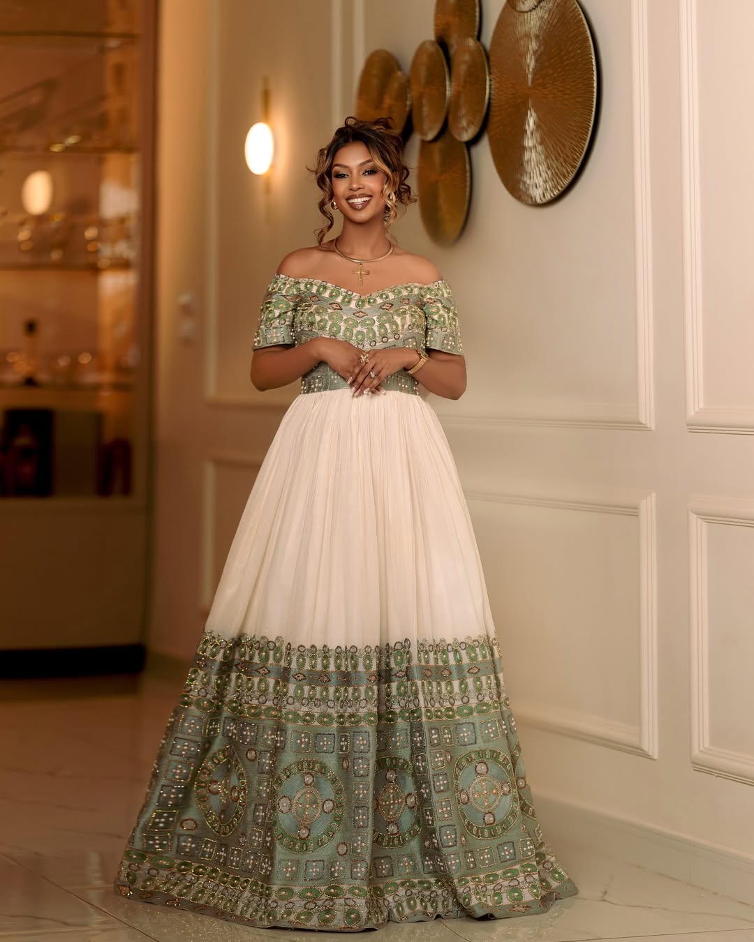 Woman wearing a traditional off-shoulder dress with green and white patterns in an elegant indoor setting.