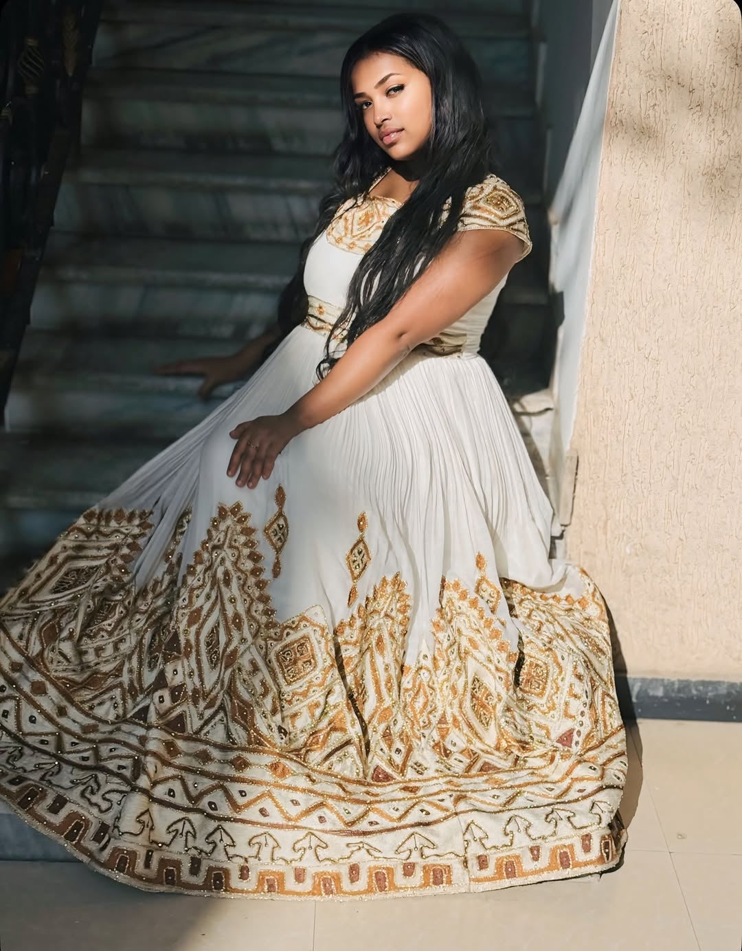 Woman in a white and gold embroidered Ethiopian traditional dress sitting on steps.