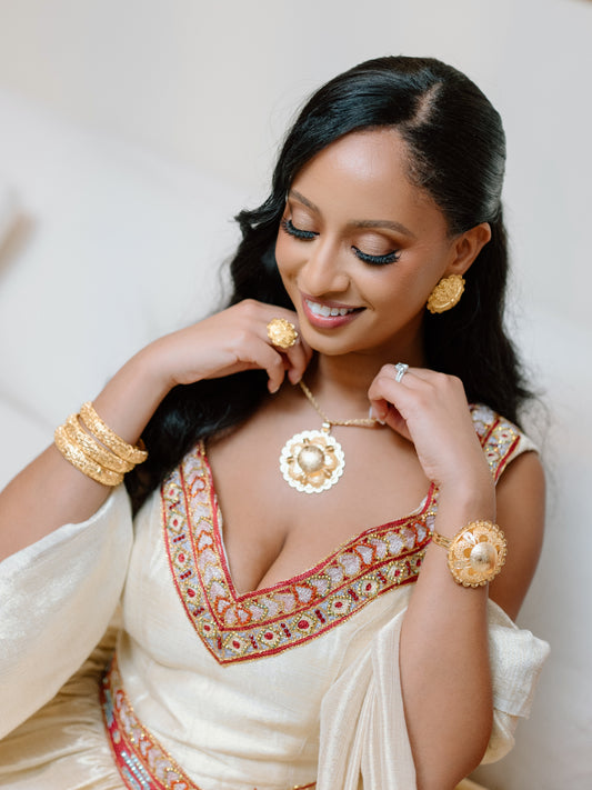 A woman wearing a cream-colored traditional ball Ethiopian national dresses with intricate red, gold, and blue geometric patterns sits on a white sofa in a living room setting.