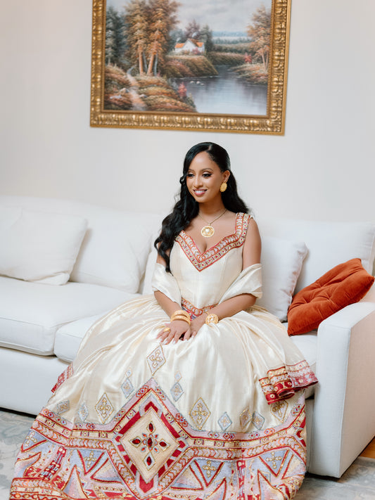 A woman wearing a cream-colored traditional ball Ethiopian national dresses with intricate red, gold, and blue geometric patterns sits on a white sofa in a living room setting.