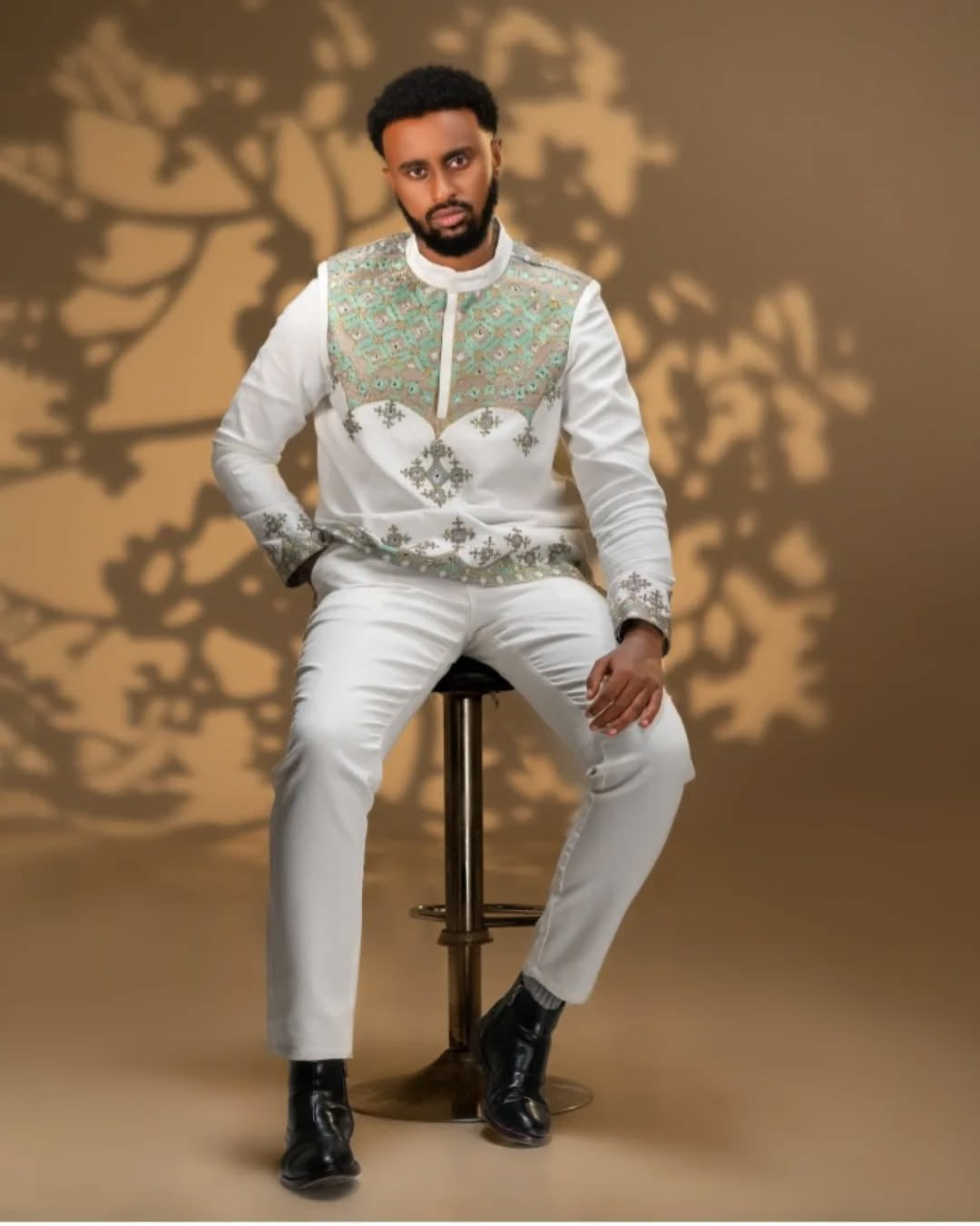 Man sitting on a chair wearing an embroidered Men’s Traditional Shirt and white pants against a brown background