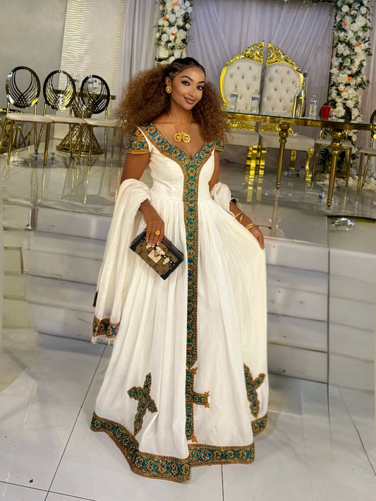 A woman wearing a white traditional Ethiopian national dress with green and gold embroidery, holding a black clutch bag, standing in a decorated event hall with white steps and gold accents.