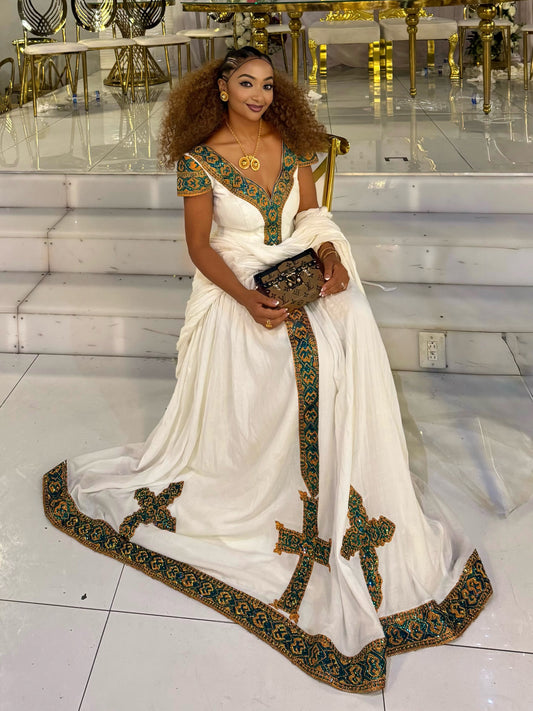 A woman wearing a white traditional Ethiopian national dress with ornate green and gold embroidered borders and a matching embroidered blouse, seated on white steps in a decorated interior.